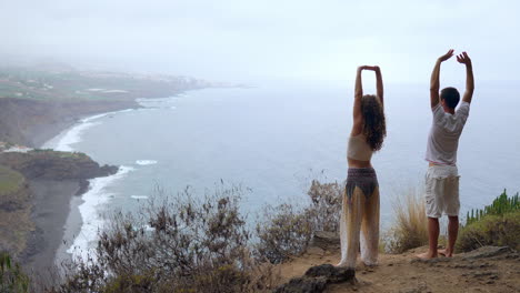 Atop-a-cliff,-a-man-and-woman-lift-their-hands,-inhaling-the-ocean-breeze-as-they-engage-in-yoga,-embracing-the-calming-atmosphere